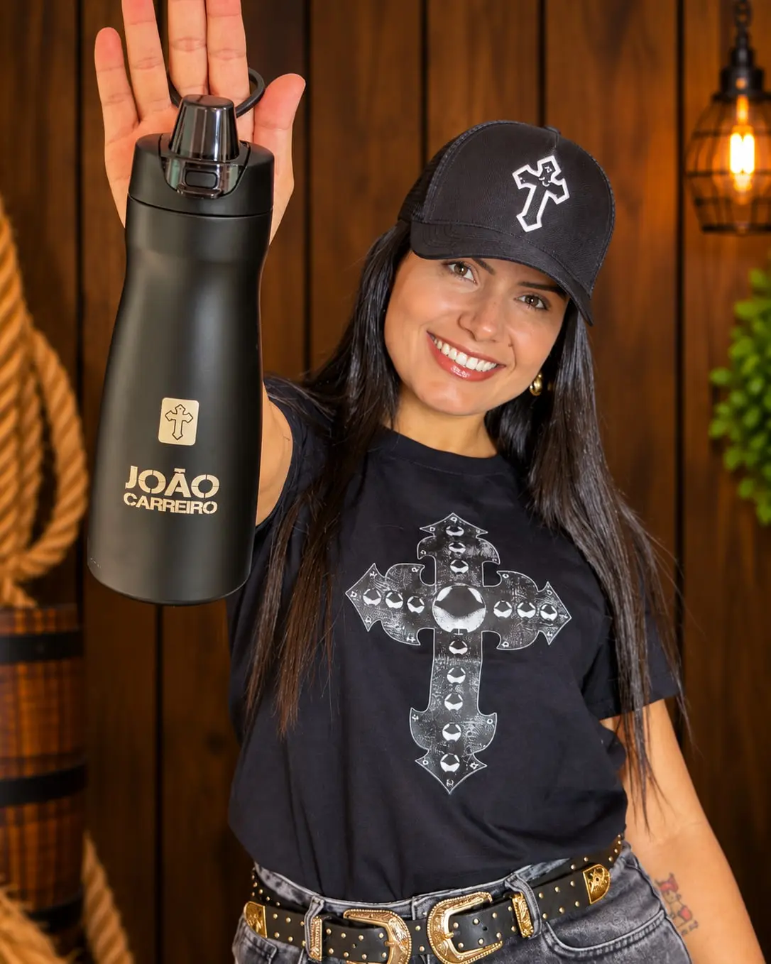 Modelo sorrindo, de boné preto, com a Camiseta Cruz do Chapéu, mostrando a garrafa João Carreiro